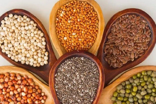 A variety of seeds and grains in wooden spoons, including white quinoa, yellow lentils, flax seeds, chia seeds, sorghum, and mung beans, arranged in a circular pattern.