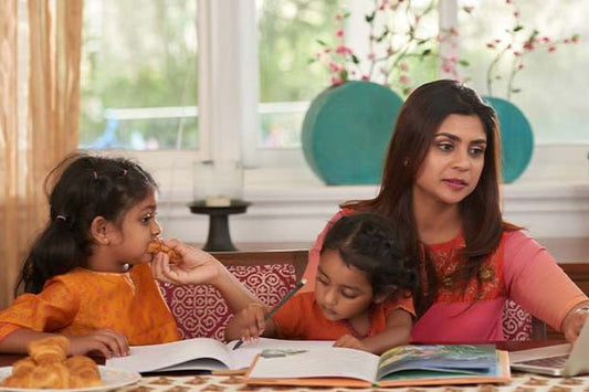 A woman seated at a table with two young girls. One girl is eating while the other looks at a book. The woman appears to be looking at something off-camera.